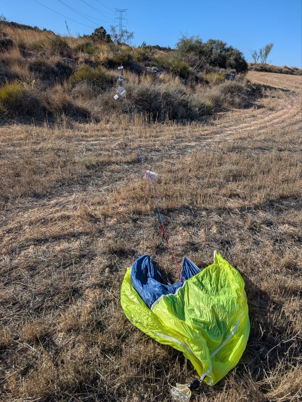Paracaídas amarillo y azul extendido sobre el rastrojo, con la línea de cápsulas de Servet XII tendida detrás